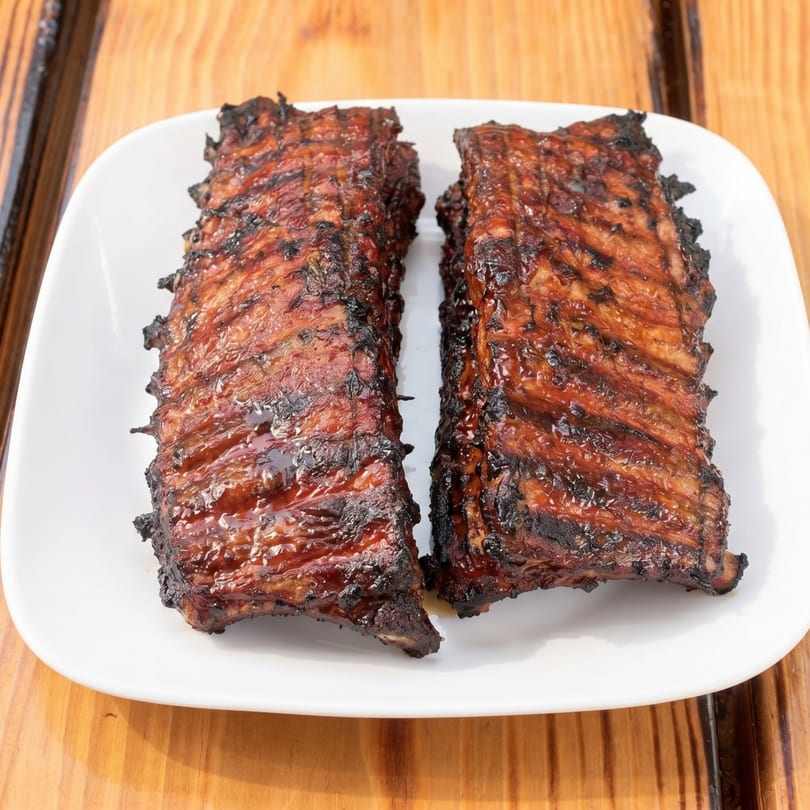 Two racks of glistening, charred BBQ ribs on a white plate, ready to be served.