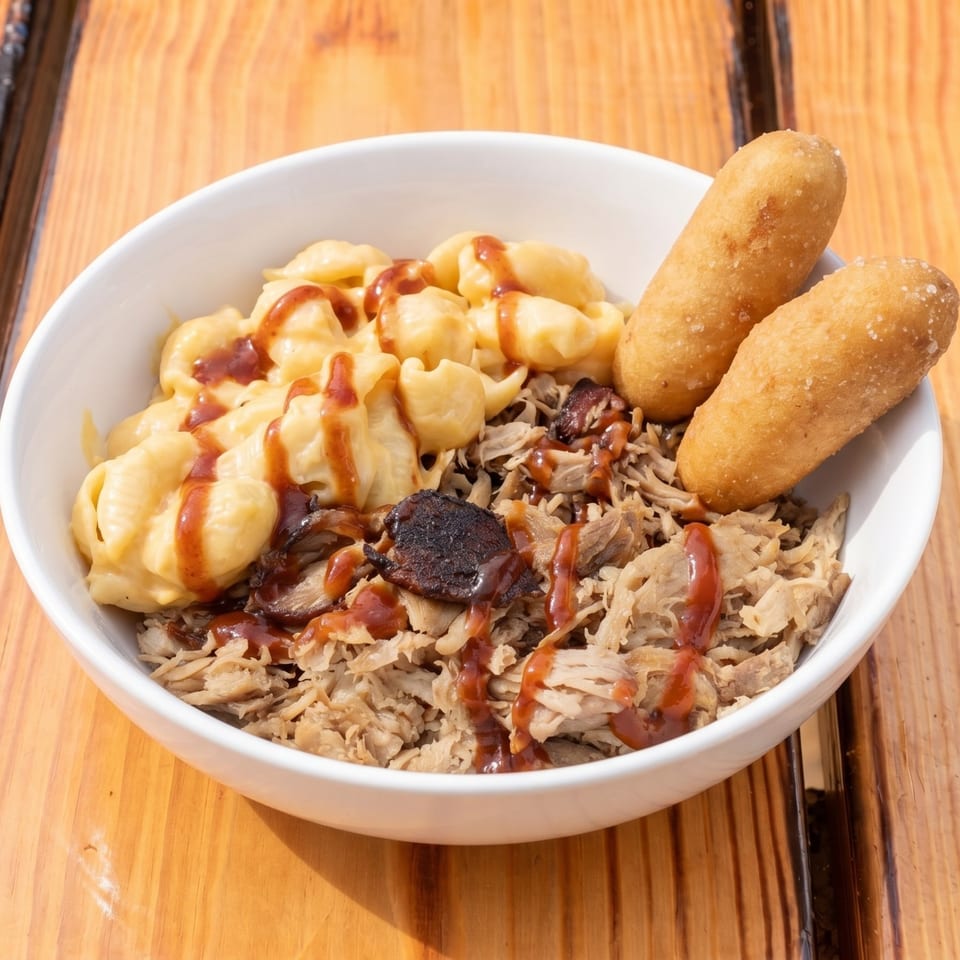 A white bowl filled with macaroni and cheese, pulled pork, and two corn dog bites, drizzled with BBQ sauce.