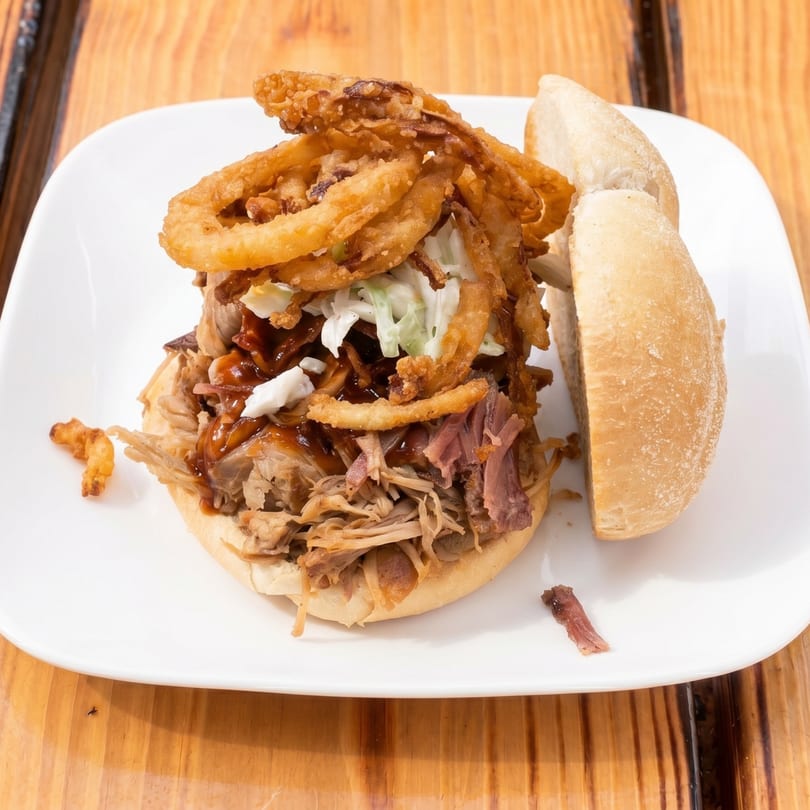 Pulled pork sandwich topped with coleslaw, crispy fried onions, and barbecue sauce on a white plate.
